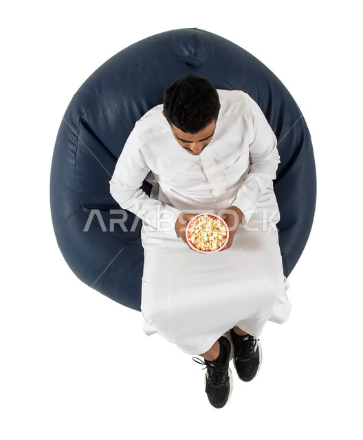 التفاعل والإندماج، بورتريه علوي عامودي لرجل عربي سعودي خليجي يرتدي الزي السعودي يجلس على كرسي يقوم بمشاهدة شيء ما  يحمل بيده علبة الفوشار , الاستمتاع بتناول الفوشار ،خلفية بيضاء