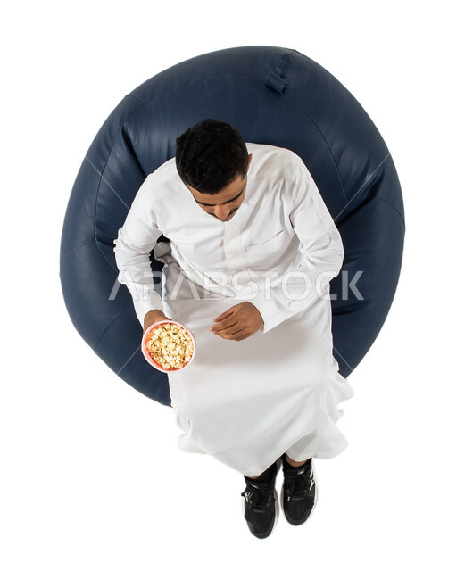 الاستمتاع بتناول الفوشار ، بورتريه علوي عامودي لرجل عربي سعودي خليجي يرتدي الزي السعودي يجلس على كرسي يقوم بمشاهدة شيء ما  يحمل بيده علبة الفوشار , خلفية بيضاء