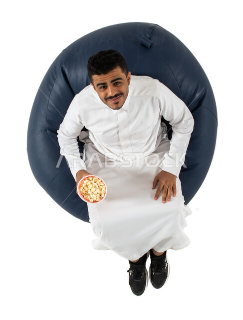 الفوز والإنتصار, بورتريه علوي عامودي لرجل عربي سعودي خليجي يرتدي الزي السعودي يجلس على كرسي يقوم بالتشجيع, يحمل بيده علبة الفوشار ، إيماءات وجه تدل على السعاده,  خلفية بيضاء