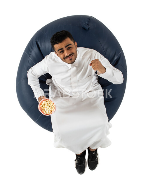 إيماءات وجه ويدين تدل على الحماس والتفاعل والإندماج والسعادة , بورتريه علوي عامودي لرجل عربي سعودي خليجي يرتدي الزي السعودي يجلس على كرسي يرفع يديه مضموم الكفين يقوم بالتشجيع ، يحمل بيده علبة الفوشار  وينظر إلى الكاميرا, خلفية بيضاء