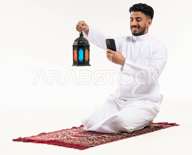 بورتريه لرجل عربي خليجي سعودي ، إلتقاط صور لفانوس رمضان بإستخدام الهاتف المحمول ، الإستعداد لقدوم شهر رمضان المبارك ، المناسبات الدينية والأعياد الإسلامية ، رمضان كريم ، إسلاميات وعبادة ، خلفية بيضاء