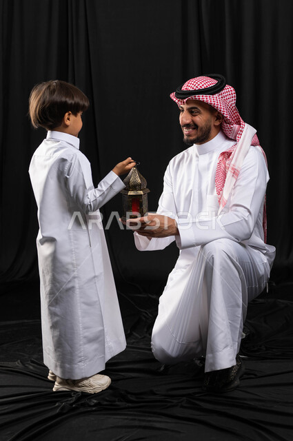 بورتريه لرجل عربي خليجي سعودي مع إبنه ، فانوس رمضان ، الإستعداد لقدوم شهر رمضان المبارك ، المناسبات الدينية والأعياد الإسلامية ، رمضان كريم ، إسلاميات وعبادة ، خلفية سوداء ، صورة بالطول الكامل