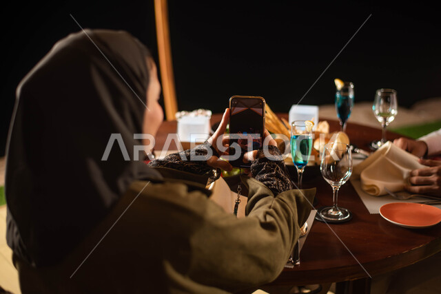 زوجان عربيان خليجيان سعوديان يستمتعان بتناول الطعام، استخدام الهاتف المحمول، التقاط صور تذكارية، التصوير من خلال الجوال، قضاء أوقات ممتعة، أجواء عائلية سعيدة، مقهى سعودي