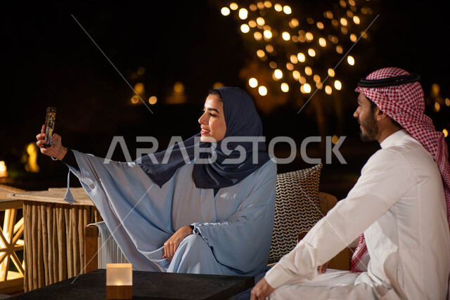 زوجان عربيان خليجيان سعوديان يستخدامان الهاتف المحمول، التقاط صورة تذكارية، صور سيلفي، الكاميرا الامامية، تقنيات حديثة، قضاء اوقات ممتعة، وناسة وتعارف، الجلوس في الهواء الطلق
