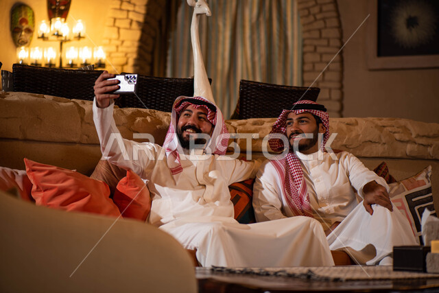 رجلان عربيان خليجيان سعوديان يرتديان الزي السعودي ويستخدامان الهاتف المحمول، التقاط صورة تذكارية، صور سيلفي، الكاميرا الامامية، صداقات شباب، تقنيات حديثة، جلسة شبابية