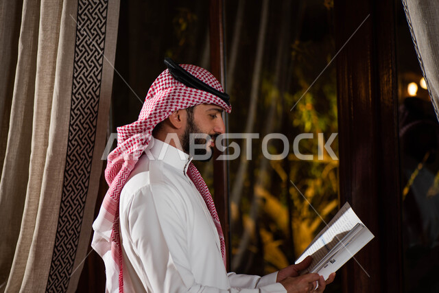 رجل عربي خليجي سعودي يحمل بيده كتاب ويقف امام النافذة، قضاء الوقت بالقراءة، التعلم والثقافة، القراءة والمعرفة، جلسة سعودية