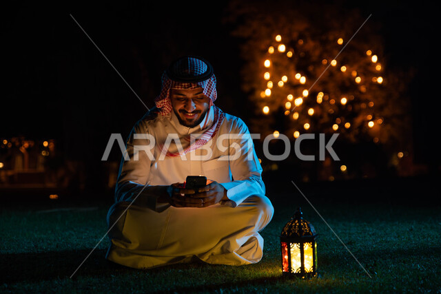 رجل عربي خليجي سعودي يستخدم الهاتف المحمول، التواصل الاجتماعي، تصفح المواقع الالكترونية، فانوس رمضان، استقبال شهر رمضان المبارك، زينة شهر رمضان، أجواء رمضانية، جلسة خارجية في الهواء الطلق