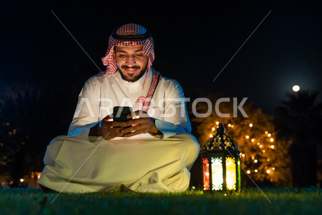 رجل عربي خليجي سعودي يستخدم الهاتف المحمول، التواصل الاجتماعي، تصفح المواقع الالكترونية، فانوس رمضان، استقبال شهر رمضان المبارك، زينة شهر رمضان، أجواء رمضانية، جلسة خارجية في الهواء الطلق
