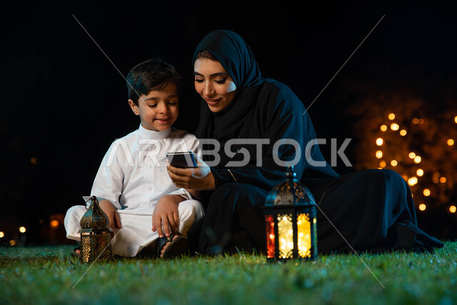 امرأة عربية خليجية سعودية وابنها، استخدام الهاتف المحمول، مشاهدة البرامج الرمضانية، تصفح المواقع الالكترونية، فوانيس رمضان، استقبال شهر رمضان المبارك، زينة شهر رمضان، أجواء رمضانية، جلسة خارجية في الهواء الطلق