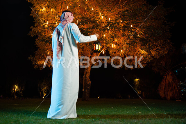 صورة من الخلف لرجل عربي خليجي سعودي يحمل بيده فانوس مضيء، استقبال شهر رمضان المبارك، المناسبات الدينية والأعياد الإسلامية، فانوس رمضان، زينة شهر رمضان، أجواء رمضانية
