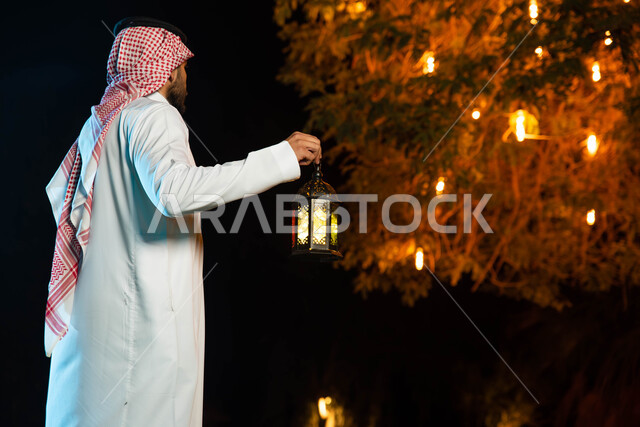 صورة من الخلف لرجل عربي خليجي سعودي يحمل بيده فانوس مضيء، استقبال شهر رمضان المبارك، المناسبات الدينية والأعياد الإسلامية، فانوس رمضان، زينة شهر رمضان، أجواء رمضانية
