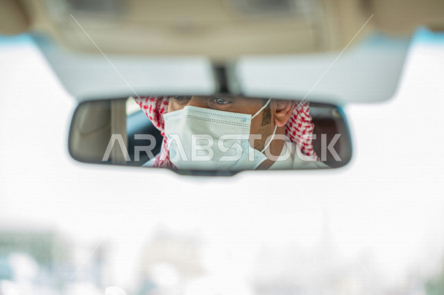 انعكاس صورة سائق شاب عربي سعودي خليجي يرتدي الكمامة في المرآه الرؤية الخلفية للسيارة ، القيادة في السعودية