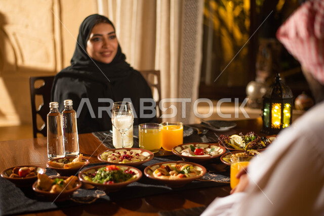 زوجان عربيان خليجيان سعوديان على مائدة الافطار بشهر رمضان، سفرة مليئة باصناف من المأكولات اللذيذة، تناول وجبة الافطار، ايماءات وجه سعيدة، فانوس رمضان، استقبال شهر رمضان المبارك، أجواء رمضانية