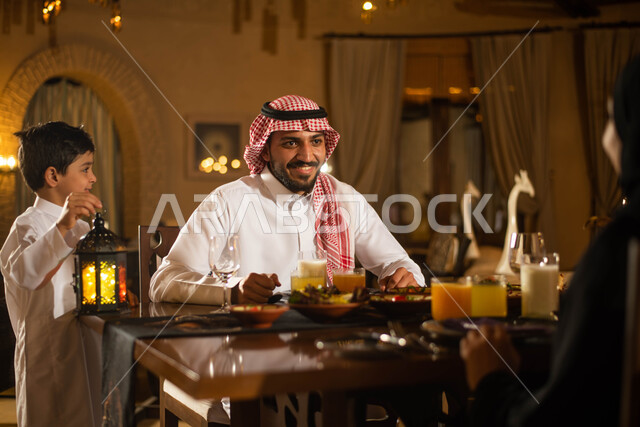 عائلة عربية خليجية سعودية على مائدة الافطار بشهر رمضان، سفرة مليئة باصناف من المأكولات اللذيذة، طفل يحمل فانوس رمضان، ايماءات وجه سعيدة، استقبال شهر رمضان المبارك، أجواء رمضانية
