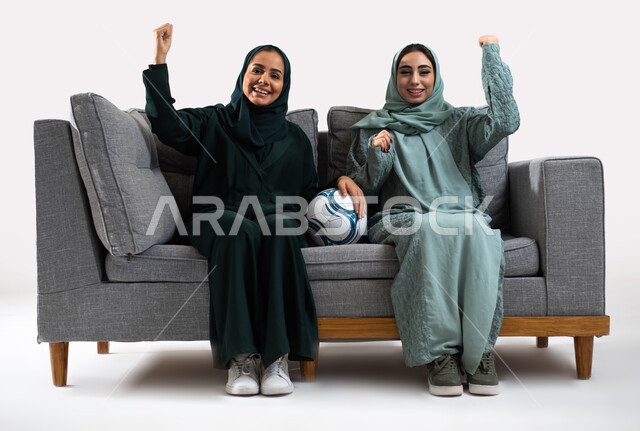 متابعة مباريات كرة القدم ،النظر إلى الكاميرا وإيماءات تدل على الفرح والسعادة لفوز الفريق ،  بورتريه لصديقتان عربيتان سعوديتان خليجيتان مبتسمتان ترتديان العباءه الخليجية ، أنديه وتشجيع ، خلفية بيضاء