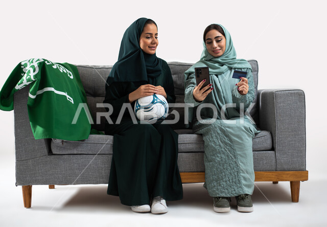 Using a bank card to book tickets to attend football matches, passion for football matches, a portrait of two Saudi Arabian Gulf women wearing the Gulf abaya, clubs and encouragement, a white background