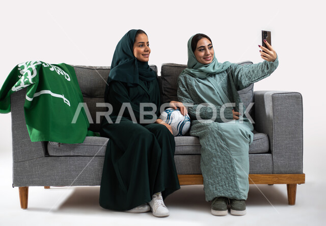 Taking selfies via the mobile device, the passion for football matches, a portrait of two smiling Saudi Arabian Gulf female friends wearing the Gulf abaya, encouraging the Saudi Al-Ahly team and the Saudi national team, clubs and encouragement, a white ba