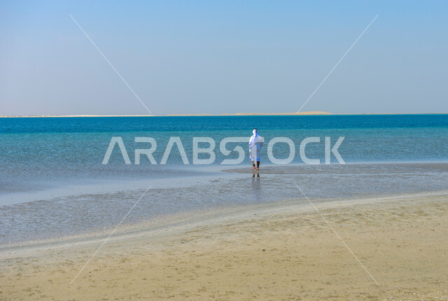 صورة من الخلف لرجل عربي خليجي قطري يقف على شاطئ البحر في منطقة خور العديد بقطر، مياه البحر الزرقاء، شاطئ البحر، رمال البحر الناعمة، مناظر طبيعية خلابة، خلفية الطبيعة