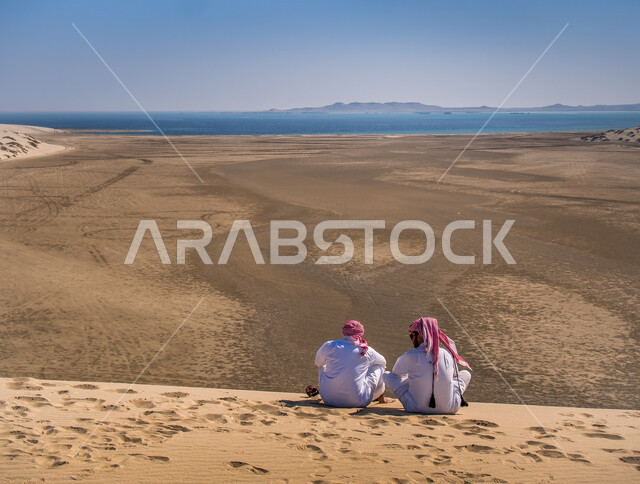 A back image of two Qatari Arab Gulf men sitting on a sandy hill overlooking the sea of Khor Al Adaid in Qatar, seashore, Khor Al Adaid area, blue sea water, soft sand, stunning landscapes, nature background