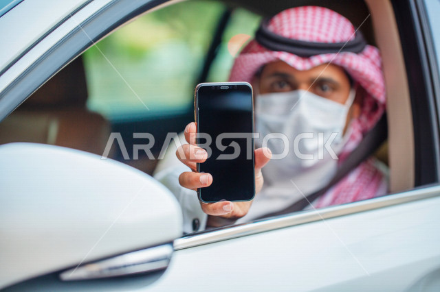 رجل عربي خليجي سعودي داخل السيارة يرتدي الكمامة للوقاية من فايروس كورونا ،  سائق يحمل بيده الهاتف المحمول ،عرض تطبيق خدمة توصيل الركاب ، تحديد الموقع ،  عرض خاصية التتبع ، التاكسي الاخضر في السعودية