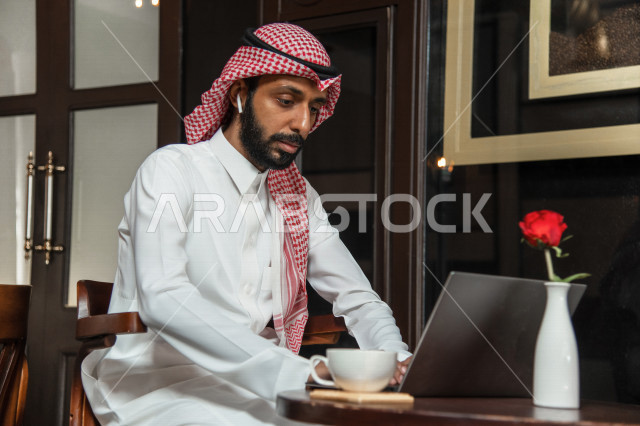 رجل أعمال عربي سعودي خليجي يجلس في المقهى, إجتماع عمل في خارج الشركه, انجهاز مهام العمل عبر جهاز الحاسوب, إدارة الأعمال عن بعد, الإستمتاع بشرب القهوه في متجر القهوه, مقاهي في المملكه العربيه السعوديه