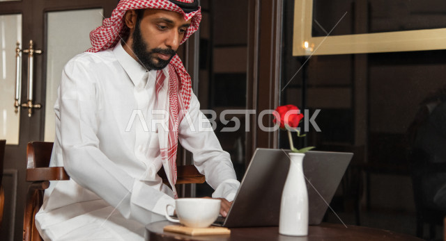 A Saudi Arabian Gulf businessman sitting in a coffee shop, working meeting outside the company, completing work tasks via a computer, managing business remotely, enjoying drinking coffee in a coffee shop, cafes in the Kingdom of Saudi Arabia