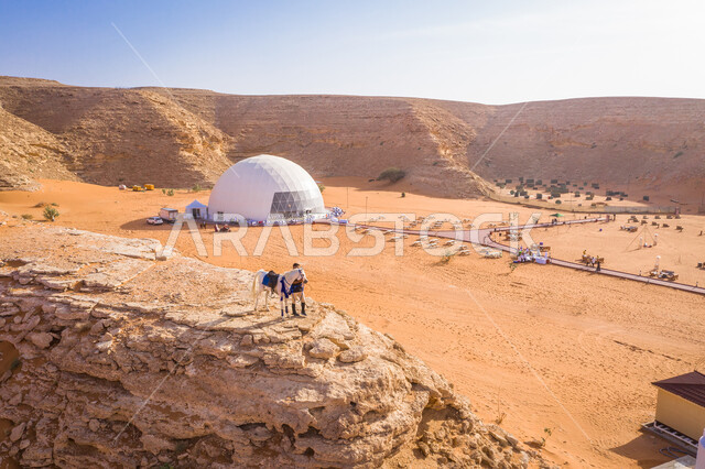 شاب عربي خليجي سعودي يقف بجانب حصان عربي اصيل فوق جبل صخري، خيمة قبة الدائرية نصف الشفافة في مخيم نوكس بصحراء المملكة العربية السعودية، الجبال والمرتفعات، الكثبان الرملية، الطبيعة الصحراوية