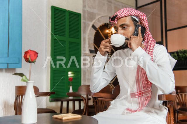 رجل عربي سعودي خليجي يجلس في المقهى, انجهاز مهام العمل عبر الهاتف المحمول, إدارة الأعمال عن بعد, دردشه ومحادثات هاتفيه, الإستمتاع بشرب القهوه في متجر القهوه, مقاهي في المملكه العربيه السعوديه