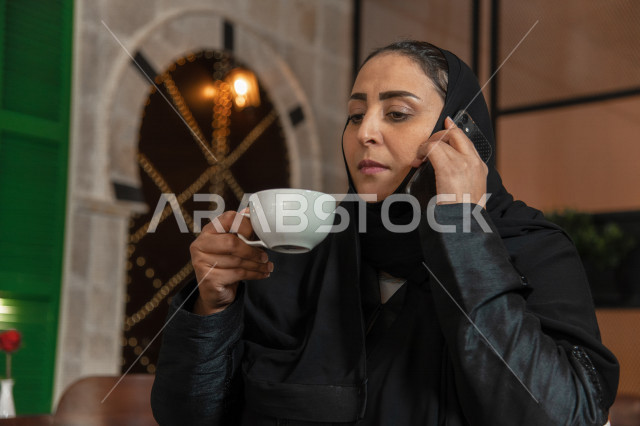 إمرأه عربيه سعوديه خليجيه تجلس في المقهى, آداء العمل عبر الهاتف المحمول, إدارة الأعمال عن بعد, دردشه ومحادثات هاتفيه, الإستمتاع بشرب القهوه في متجر القهوه, مقاهي في المملكه العربيه السعوديه