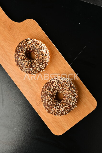 صورة من الاعلى لقطع من حلوى الدونات المزينة بالشوكلاتة على لوح خشبي، حلويات وسكريات، قطع الحلوى الشهية، حلوى الدونات، خلفية سوداء