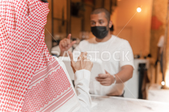منظر من الخلف لرجل عربي سعودي خليجي يرتدي الثوب والشماغ السعودي, طلب القهوه من المقهى, الدفع عند الكاشير, الدفع الإلكتروني ببطاقة الصراف, إلتزام البائع بلبس الكمامه, مقاهي في المملكه العربيه السعوديه