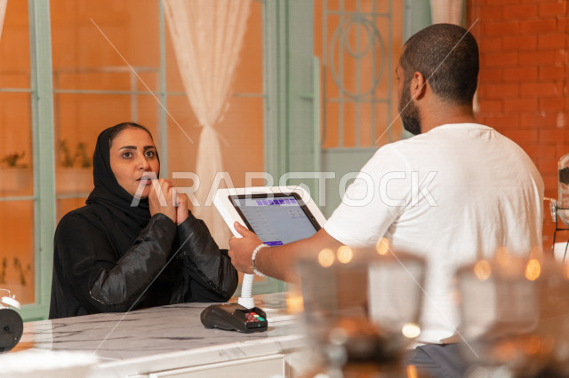 إمرأه عربيه سعوديه خليجيه ترتدي الحجاب والعباءه في متجر القهوه, طلب القهوه من المقهى, الدفع عند الكاشير , الدفع الإلكتروني ببطاقة الصراف, مقاهي في المملكه العربيه السعوديه