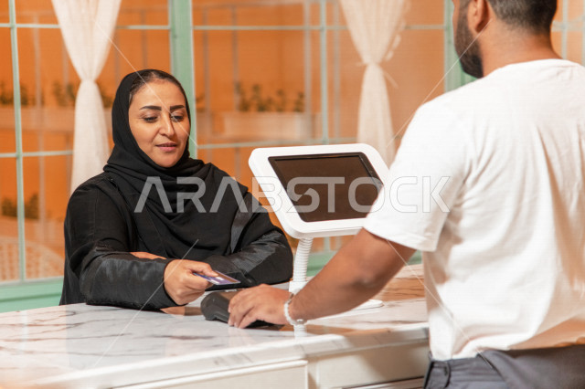 إمرأه عربيه سعوديه خليجيه ترتدي الحجاب والعباءه في متجر القهوه, طلب القهوه من المقهى, الدفع عند الكاشير , الدفع الإلكتروني ببطاقة الصراف, إيماءات وجه تدل على السعاده, مقاهي في المملكه العربيه السعوديه