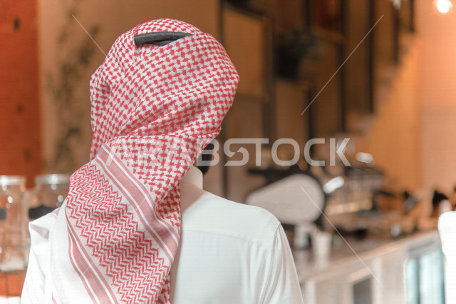 منظر من الخلف لرجل عربي سعودي خليجي يرتدي الثوب والشماغ السعودي, طلب القهوه من المقهى, إستلام القهوه, الإستمتاع بشرب القهوه في المقهى, الدفع عند الكاشير, مقاهي في المملكه العربيه السعوديه