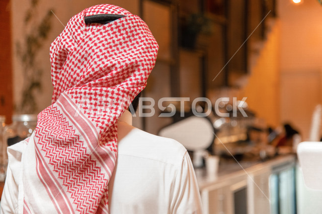 منظر من الخلف لرجل عربي سعودي خليجي يرتدي الثوب والشماغ السعودي, طلب القهوه من المقهى, إستلام القهوه, الإستمتاع بشرب القهوه في المقهى, الدفع عند الكاشير, مقاهي في المملكه العربيه السعوديه