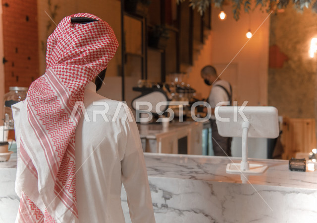 منظر من الخلف لرجل عربي سعودي خليجي يرتدي الثوب والشماغ السعودي, طلب القهوه من المقهى, إستلام القهوه, الإستمتاع بشرب القهوه في المقهى, الدفع عند الكاشير, مقاهي في المملكه العربيه السعوديه
