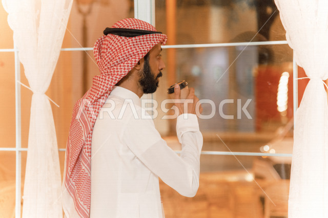 منظر جانبي لرجل عربي سعودي خليجي يرتدي الثوب والشماغ السعودي, طلب القهوه من المقهى, إستلام القهوه, الإستمتاع بشرب القهوه في المقهى, مقاهي في المملكه العربيه السعوديه
