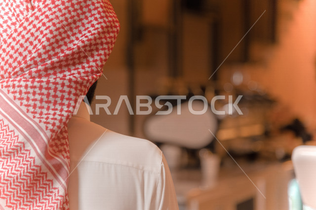 منظر من الخلف لرجل عربي سعودي خليجي يرتدي الثوب والشماغ السعودي, طلب القهوه من المقهى, إستلام القهوه, الإستمتاع بشرب القهوه في المقهى, الدفع عند الكاشير, مقاهي في المملكه العربيه السعوديه