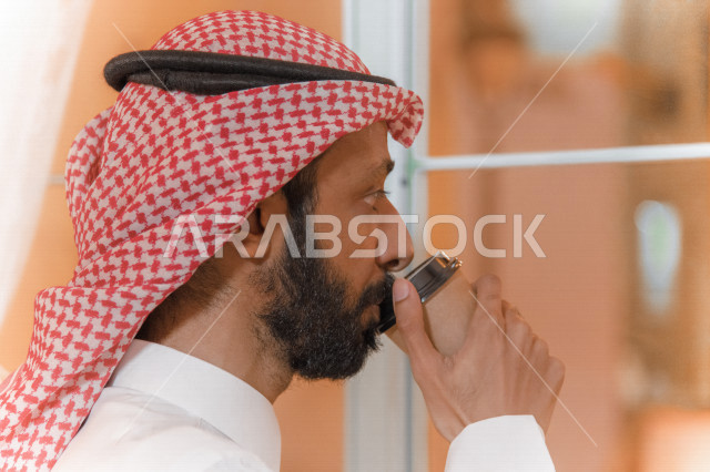 منظر جانبي لرجل عربي سعودي خليجي يرتدي الثوب والشماغ السعودي, طلب القهوه من المقهى, إستلام القهوه, الإستمتاع بشرب القهوه في المقهى, مقاهي في المملكه العربيه السعوديه