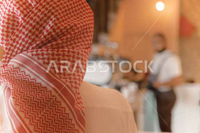 منظر من الخلف لرجل عربي سعودي خليجي يرتدي الثوب والشماغ السعودي, طلب القهوه من المقهى, إستلام القهوه, الإستمتاع بشرب القهوه في المقهى, الدفع عند الكاشير, مقاهي في المملكه العربيه السعوديه
