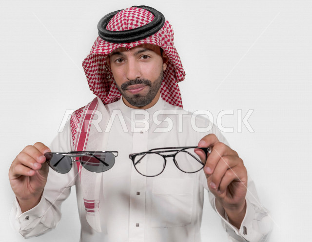 بورتريه لرجل عربي خليجي سعودي يرتدي الزي السعودي التقليدي و يحمل بيده نظارة طبية و باليد الاخرى نظارة شمسية بإيماءات و تعابير وجه تدل على الابتسامة ، المقارنة بين النظارات الطبية و الشمسية ، خلفية بيضاء