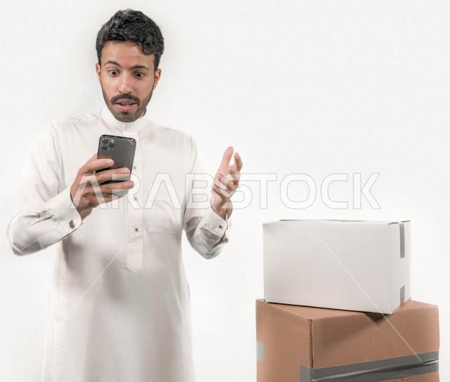 بورتريه لشاب عربي خليجي سعودي يقوم بإرسال الطلبات اونلاين باستخدام الهاتف المحمول بإيماءات و تعابير تدل على الدهشة ، طلبات اونلاين ، التسوق عن بعد ، خلفية بيضاء
