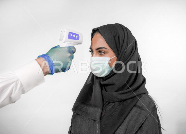 بورتريه لشابة عربية خليجية سعودية ترتدي الكمامة ، تقوم بفحص درجة الحرارة بإستخدام ميزان الحرارة الرقمي لقياس درجة الحرارة ، اتباع إجراءات الوقاية و السلامة من فايروس كورونا ، خلفية بيضاء