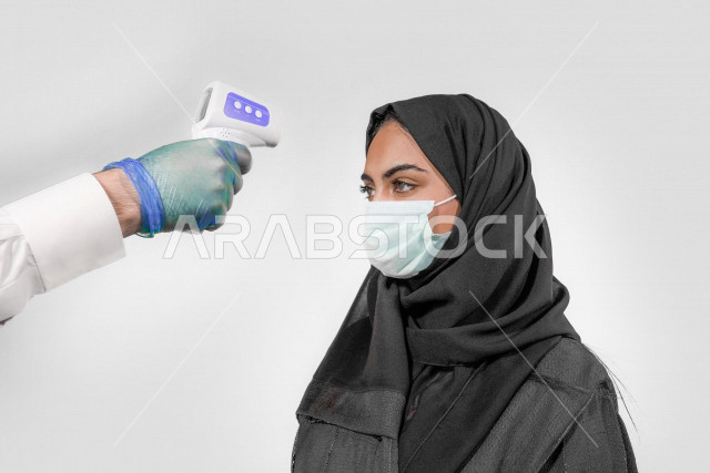 بورتريه لشابة عربية خليجية سعودية ترتدي الكمامة ، تقوم بفحص درجة الحرارة بإستخدام ميزان الحرارة الرقمي لقياس درجة الحرارة ، اتباع إجراءات الوقاية و السلامة من فايروس كورونا ، خلفية بيضاء