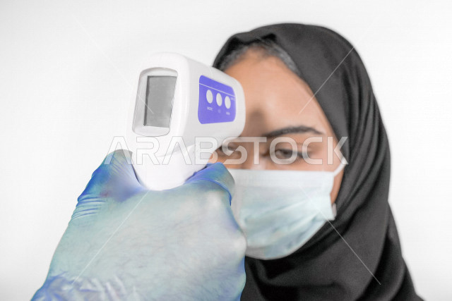 صورة مقربة لشابة عربية خليجية سعودية ترتدي الكمامة ، تقوم بفحص درجة الحرارة بإستخدام ميزان الحرارة الرقمي لقياس درجة الحرارة ، اتباع إجراءات الوقاية و السلامة من فايروس كورونا ، خلفية بيضاء