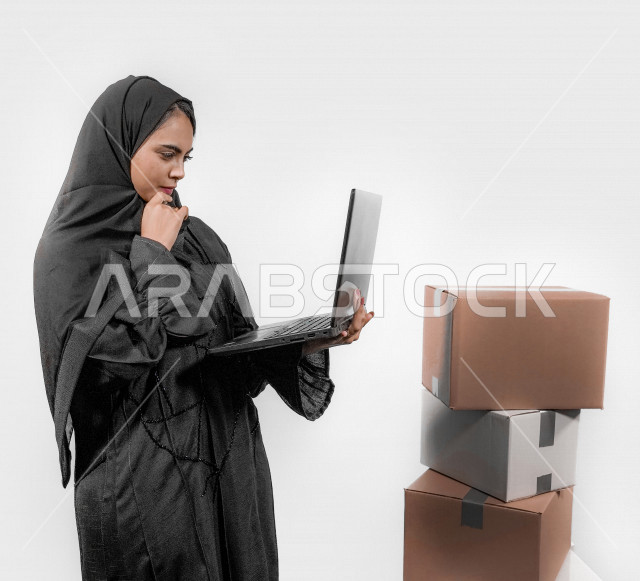 بورتريه لشابة عربية خليجية سعودية تقوم بحمل جهاز الحاسوب المحمول و استخدام تطبيقات التسوق عن بعد بإيماءات تدل على التركيز ، إرسال الطلبات اونلاين ، طلبات اونلاين ، مبيعات و مشتريات ، خلفية بيضاء