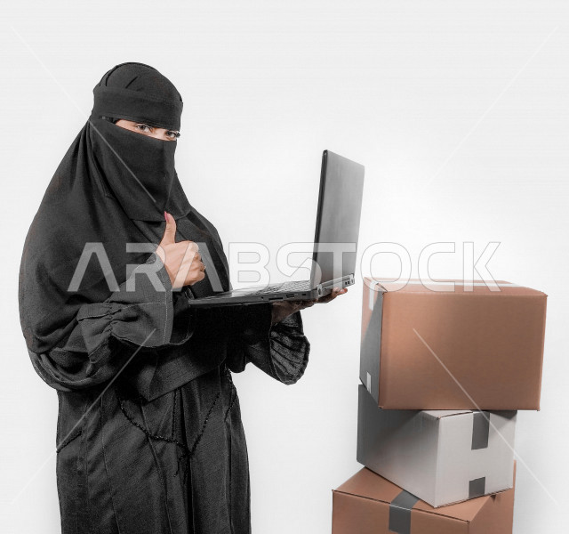 بورتريه لشابة عربية خليجية سعودية منقبة تقوم بحمل جهاز الحاسوب المحمول و استخدام تطبيقات التسوق عن بعد ، تشير بيدها بإشارة الاعجاب للعمل الممتاز ، إرسال الطلبات اونلاين ، مبيعات و مشتريات ، خلفية بيضاء