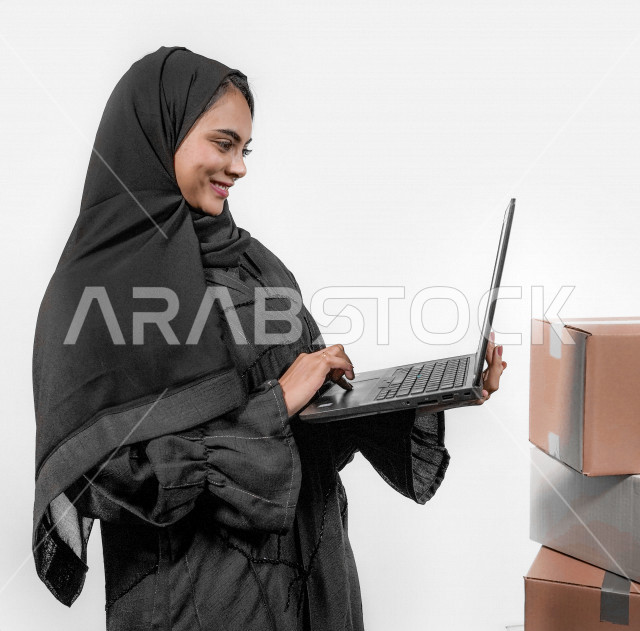 بورتريه لشابة عربية خليجية سعودية مبتسمة تقوم بحمل جهاز الحاسوب المحمول و استخدام تطبيقات التسوق عن بعد ، إرسال الطلبات اونلاين ، طلبات اونلاين ، مبيعات و مشتريات ، خلفية بيضاء