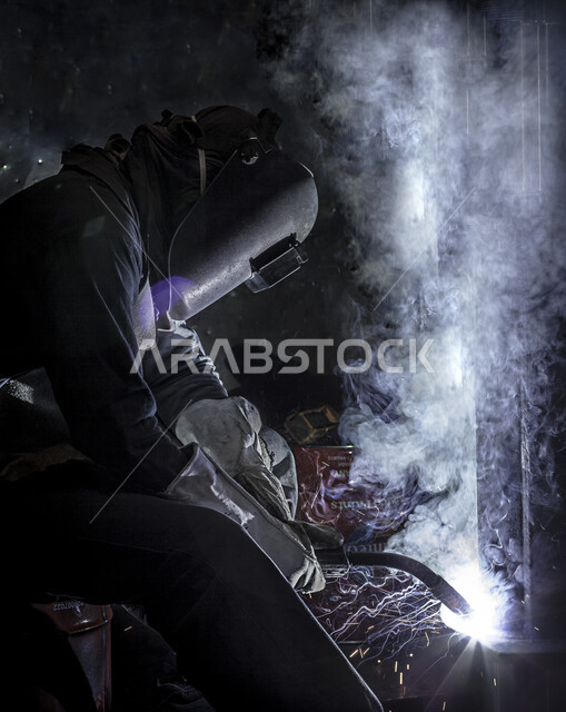 عامل عربي خليجي سعودي داخل مصنع الحديد ، ارتداء قناع الحماية في عمل لحام الحديد ، الحماية من شرارة لحام الحديد ، عملية انتاج الحديد .