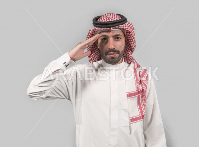 بورتريه لرجل عربي خليجي سعودي يرتدي الزي التقليدي السعودي رافعا يده اليمنى لأداء التحية العسكرية بإيماءات و تعابير تدل على الإستعداد و الإنضباط ، خلفية بيضاء